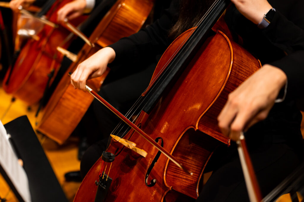 A zoomed-in image of a cello with only the body of the instrument and the musician's bow hand visible. Two cellos are similarly visible on the left-hand side of the photo, and one musician's bow hand is visible on the right-hand side of the photo.
